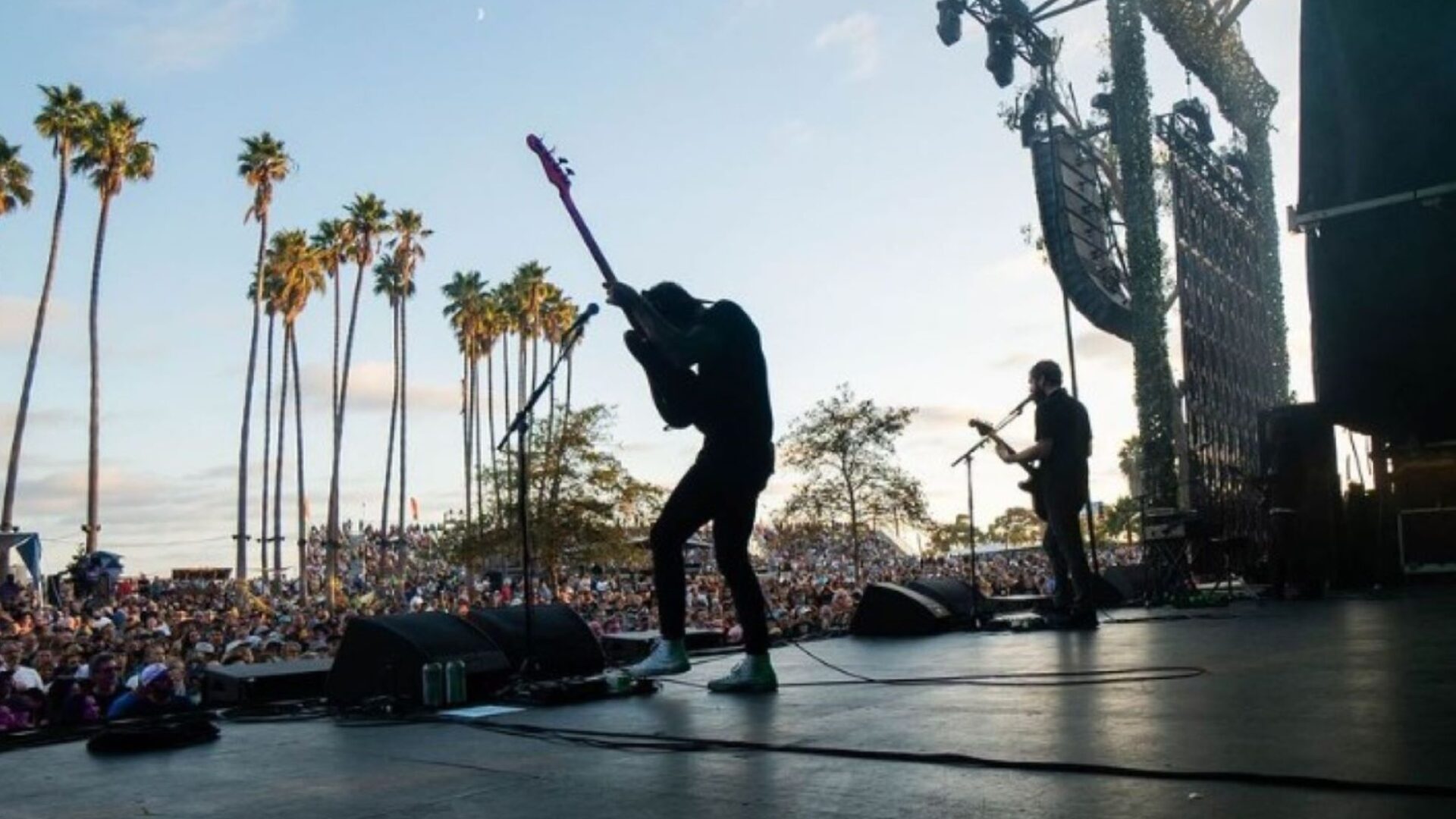 Musicians on stage playing guitar with crowd and palm trees in background