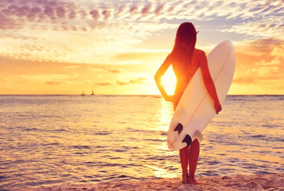 Girl with long hair standing at the shore of a beach at sunset holding a white surfboard