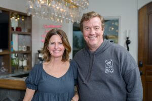 Man in a grey hooded sweatshirt standing next to a brunette woman in a blue shirt